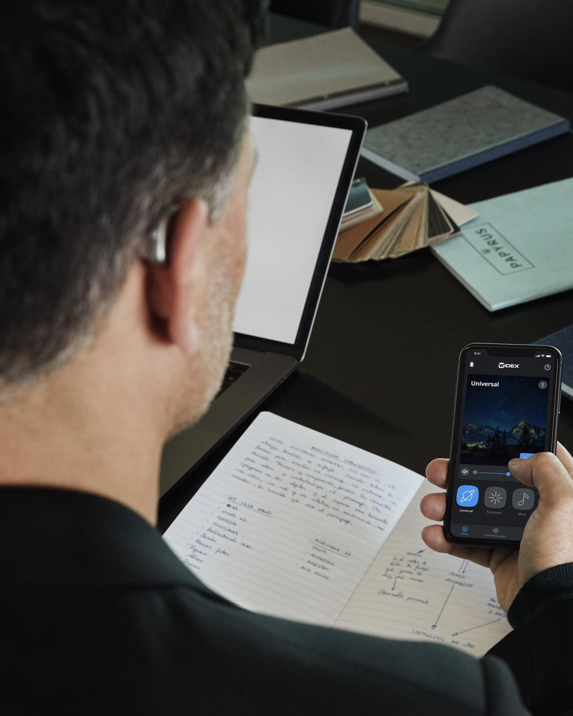 Ein Hörgeräteträger blickt auf ein Smartphone-Display, auf dem eine Hör-App angezeigt wird, während auf dem Schreibtisch ein aufgeschlagenes Notizbuch mit handschriftlichen Notizen und ein Laptop liegen.